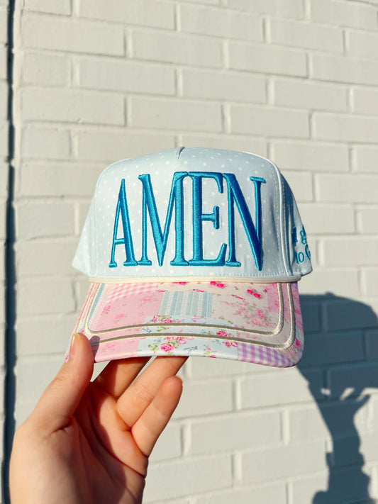 Cap with 'AMEN' text held against a white brick wall