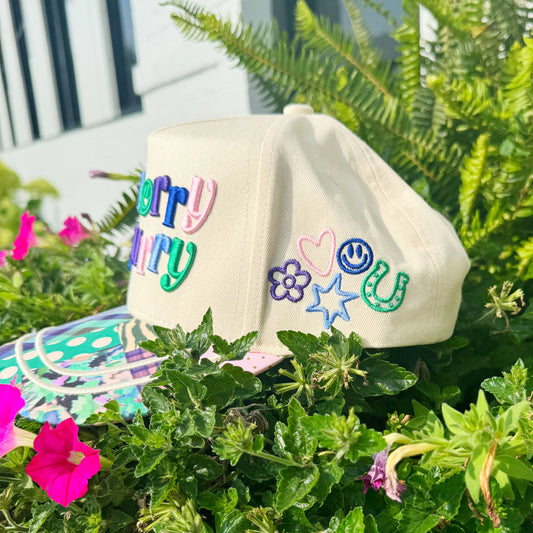Beige cap with colorful embroidered text and designs on a green plant background