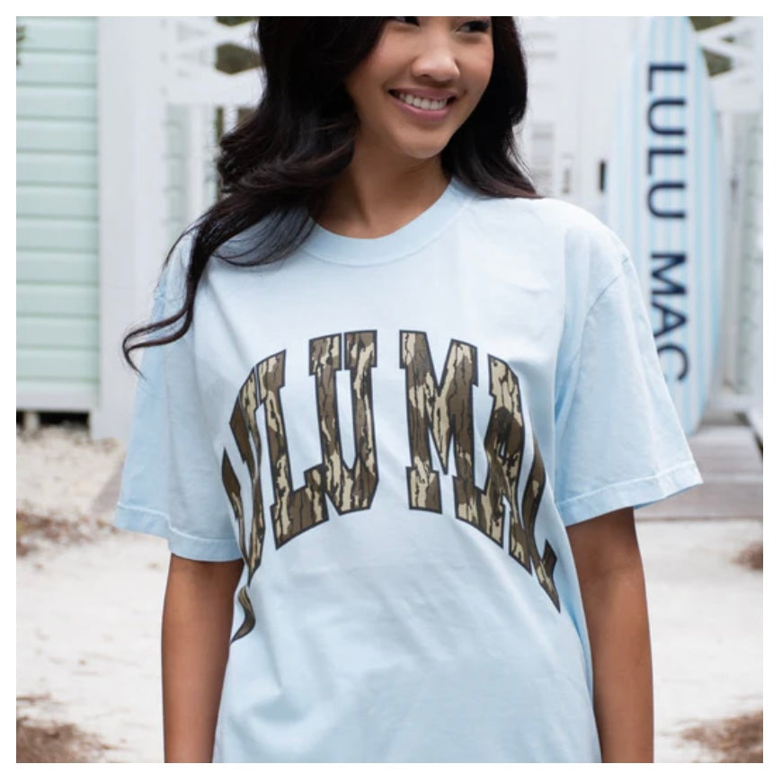 Woman wearing a light blue t-shirt with 'LULU MAC' printed on it, standing outdoors.