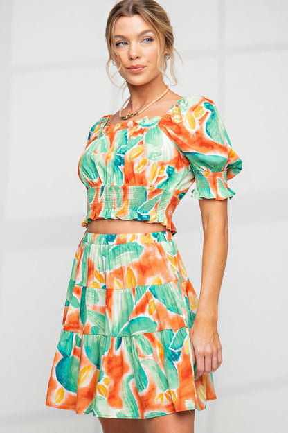 A woman wearing a two-piece outfit with a smocked crop top with puff sleeves and a matching tiered mini skirt in a vibrant tropical watercolor print.