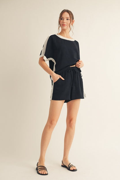 A female model wearing a black oversized short-sleeve shirt with white detailing and matching black shorts with white side stripes, paired with black sandals.